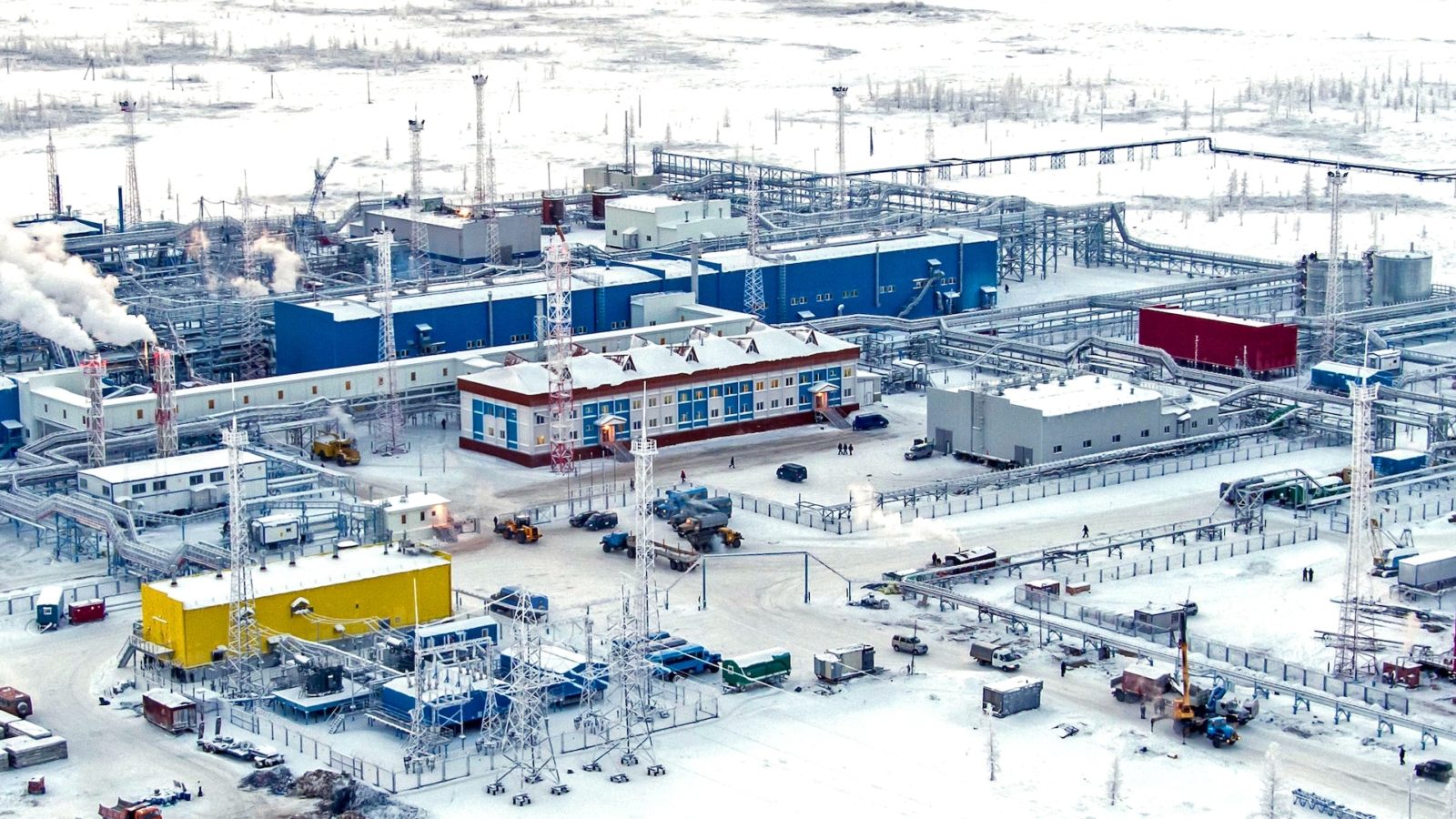 Industrial buildings on a snowy landscape with gas coming out of towers in the buildings.